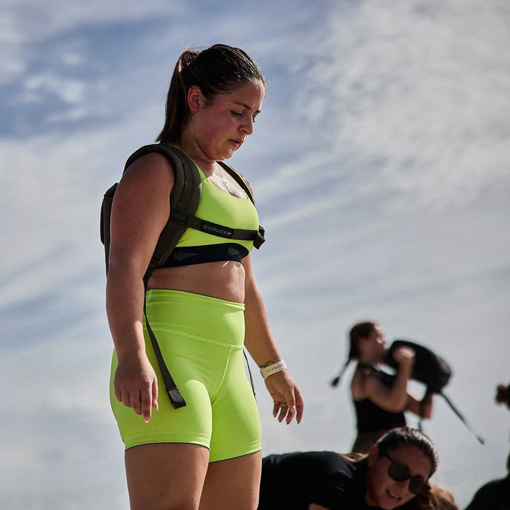 A woman wearing neon green Women's Biker Shorts made with GORUCK's ToughFlex fabric stands outdoors under a cloudy sky, carrying a weighted vest. Another person in the background adjusts their equipment. The scene suggests participation in an outdoor fitness activity, highlighting the stay-put technology that ensures everything remains secure.