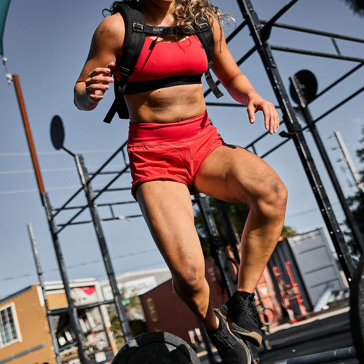 An athlete in a red outfit made from GORUCK's ToughStretch® fabric performs a box jump at an outdoor gym. The squat-proof Women’s Training Shorts - ToughStretch provide her comfort and confidence under the clear sky.