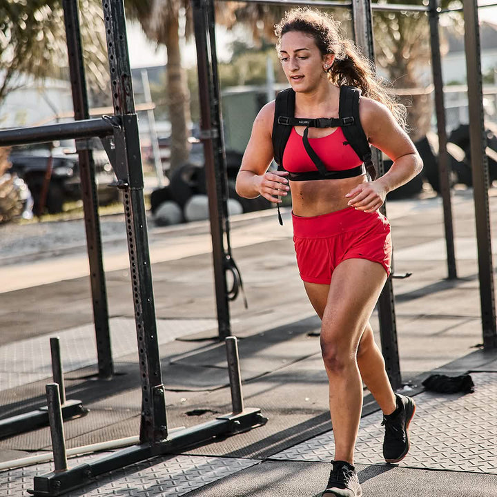 A woman in red athletic gear, including GORUCK's Women's Training Shorts made from squat-proof ToughStretch® fabric, jogs outdoors with a weighted vest, passing gym equipment.