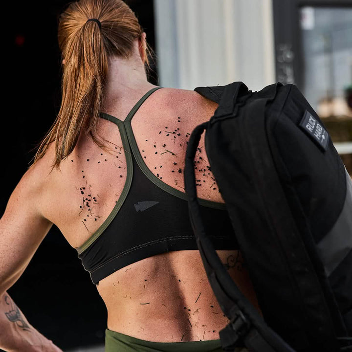 A woman with long hair in a ponytail, wearing the GORUCK Stealth Bra - ToughFlex that offers lightweight support and breathability, carries a black backpack. Small pieces of shredded rubber cling to her back as she stands outside a building, facing away from the camera.