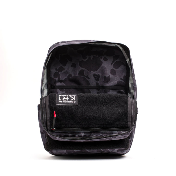 Introducing the KR1 2.0 - Kid Ruck by GORUCK: a robust children's backpack with a sleek black camouflage pattern, an accessible open front pocket, and a striking red zipper pull. It's crafted with reinforced stitching for added durability.