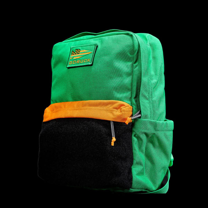 The GORUCK KR1 2.0 - Kid Ruck is a durable backpack for kids, available in green with an orange and black front pocket, and it includes reinforced stitching for added strength, presented against a black background.