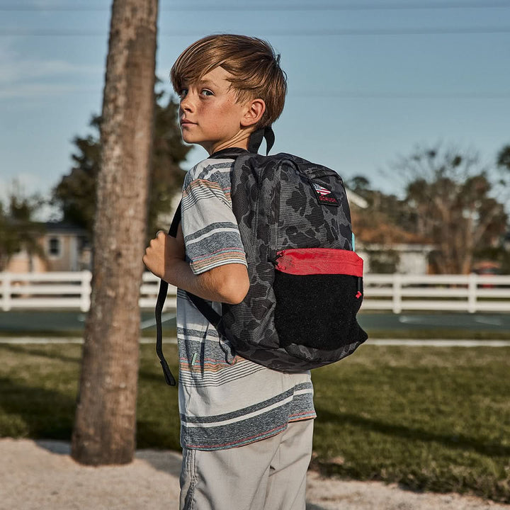 A boy wearing a striped shirt stands outdoors near trees and a fence, ready for adventure with the GORUCK KR1 2.0 - Kid Ruck on his back. Featuring reinforced stitching, this durable pack is perfect for all his escapades.