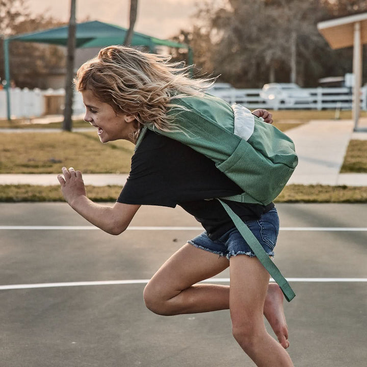 A child with a GORUCK KR1 2.0 - Kid Ruck runs joyfully on an outdoor court in casual clothes, showcasing its reinforced stitching.