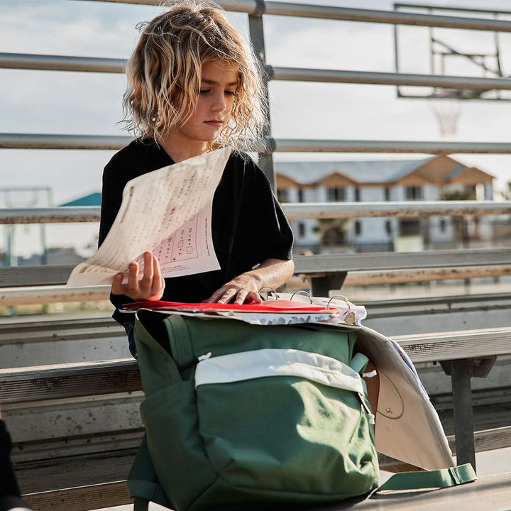 The child sits on the bleachers, holding a sheet of paper, with a GORUCK KR1 2.0 - Kid Ruck backpack featuring reinforced stitching beside them.