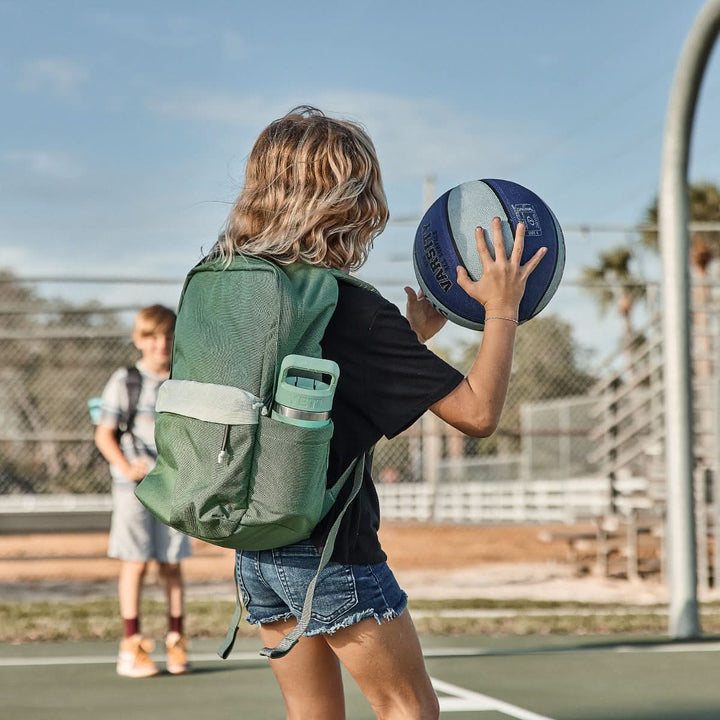 A person with a KR1 2.0 - Kid Ruck backpack by GORUCK, featuring reinforced stitching, holds a basketball on an outdoor court, while another person is visible in the background.