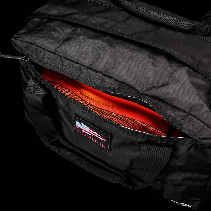 The Kit Bag - Dyneema by GORUCK, a black aviator's kit bag with durable handles and a zippered pocket, is open, showcasing an orange mesh interior. A patch featuring a red, white, and blue design with the brand name GORUCK suggests its Special Forces inspiration.