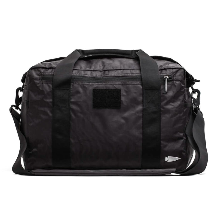 Introducing the GORUCK Kit Bag - Dyneema: A sleek, black duffel bag inspired by an Aviator’s Kit Bag. It features sturdy handles and a detachable shoulder strap, complemented by a front zipper pocket and a small black patch. Designed for minimalist style, this bag is perfect for travel or gym use.