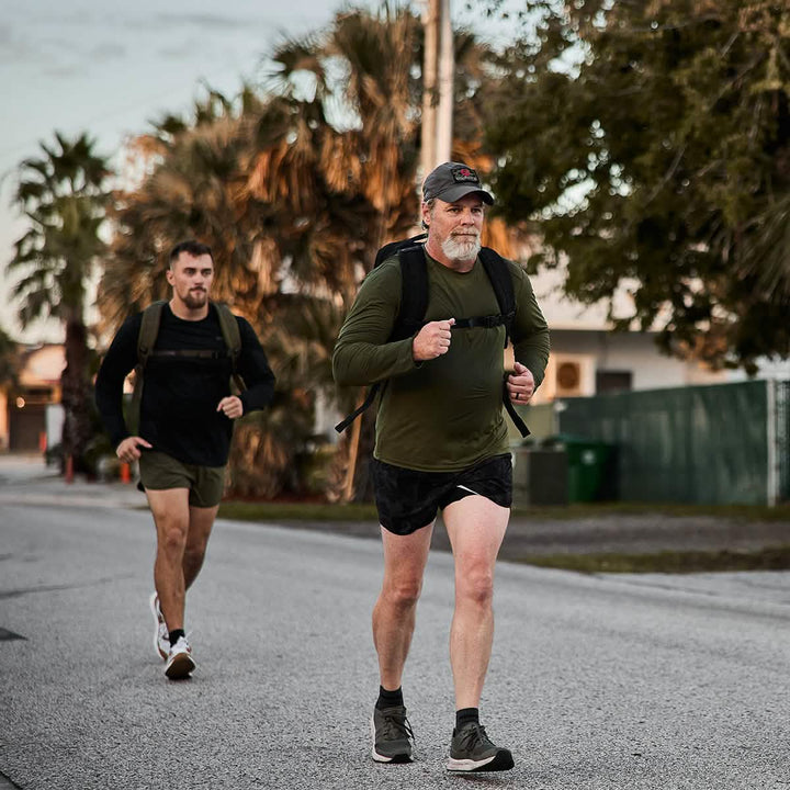 Two men dressed in athletic attire, featuring GORUCK's Men's Long Sleeve Performance Tee made with ToughMesh fabric, are walking briskly along a paved road. The outdoor scene is complemented by palm trees and a fence in the background, while they look focused and determined.