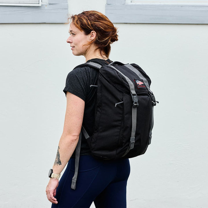 A person with a tattoo on their left forearm wears the GORUCK M23 backpack made of durable Cordura materials and a black T-shirt, standing in profile against a light-colored wall. They have short brown hair and are wearing a wristwatch.