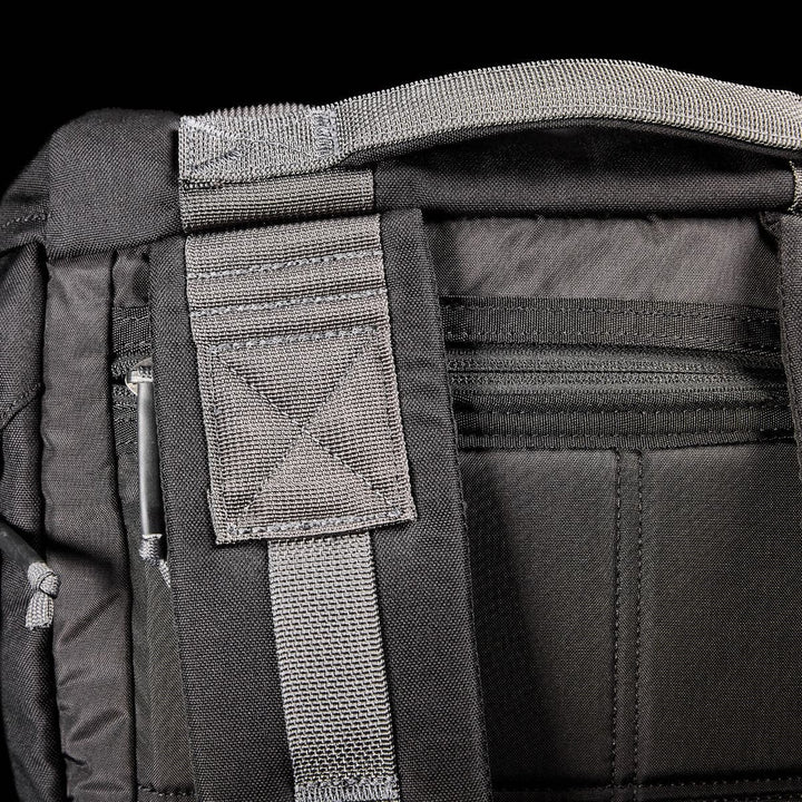 Close-up of a black GORUCK M23 - Cordura rucksack, highlighting its robust shoulder straps and padded back panel. Made from durable materials with intricate stitching, it features a partially visible zipper pocket for additional storage. The contemporary rucksack is striking against the dark background.