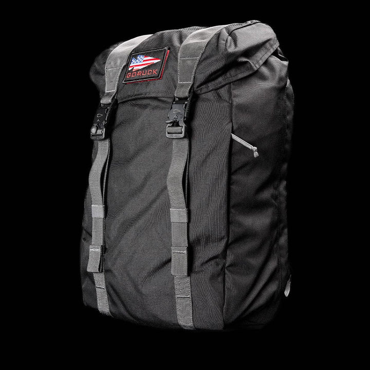 A black GORUCK M23 - Cordura rucksack showcases a rectangular shape, enhanced by a top flap with two adjustable buckle straps and a logo patch on the flap. Constructed from durable materials, it features a front zipper pocket and is set against a solid black backdrop.