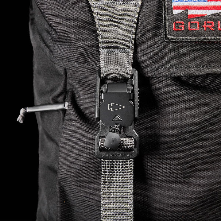 The GORUCK M23 - Cordura rucksack is showcased up close, illustrating its robust construction and secure buckle fastening of the gray strap. The strap displays a small triangular arrow symbol, and there's a partly visible patch with American flag colors in the top right corner.