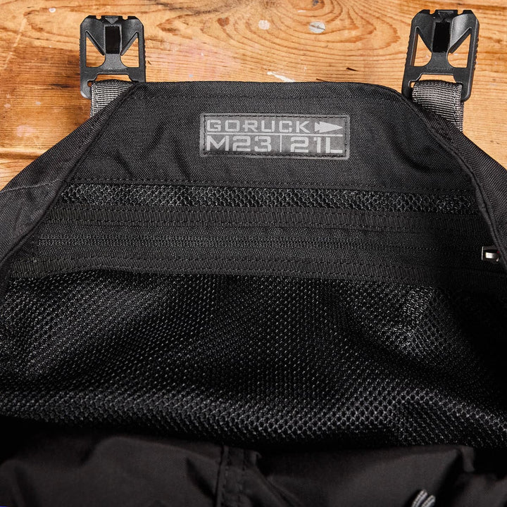 A close-up of a sleek black GORUCK M23 - Cordura backpack laid flat, displaying the label on the upper inside section. The modern rucksack's interior mesh pocket is visible, while the wooden surface beneath adds texture to the image.