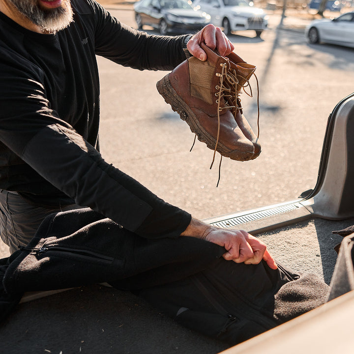 Someone carefully packs a pair of MACV-2 Mid Top boots by GORUCK, featuring a briar and coyote color with an aggressive triple compound outsole, into a bag at the back of a vehicle, ready for adventure.