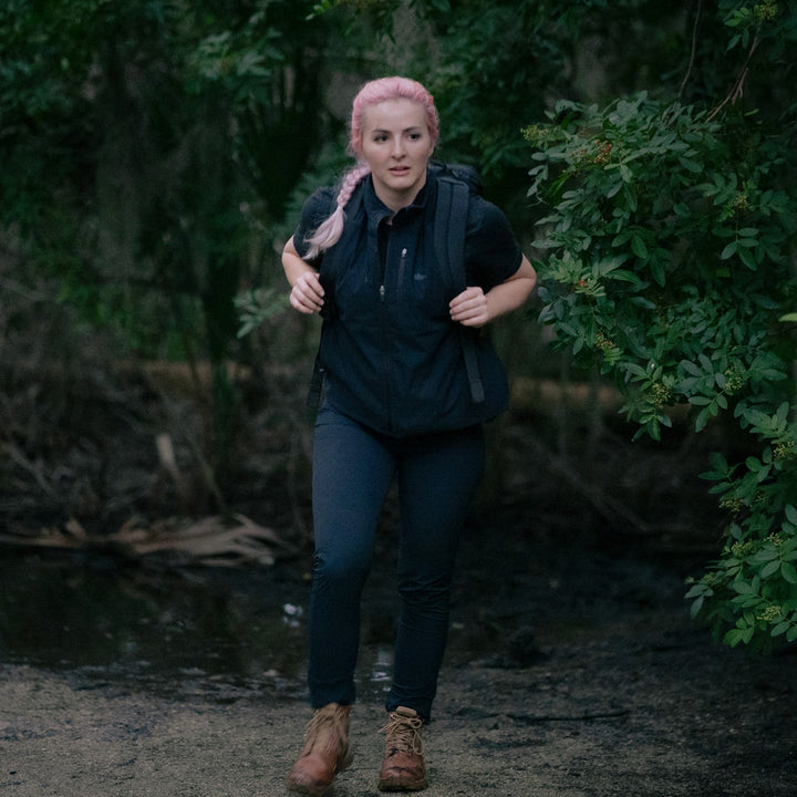 A person with pink hair hikes through a forest, wearing a black outfit and carrying a backpack, while sporting GORUCK MACV-2 - Mid Top - Briar + Coyote jungle boots featuring an aggressive triple compound outsole.