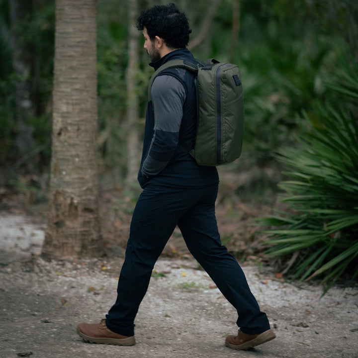 A man strolls through the forest, wearing a backpack, dark clothing, and GORUCK's MACV-2 Mid Top boots in Briar + Coyote with an aggressive triple compound outsole.