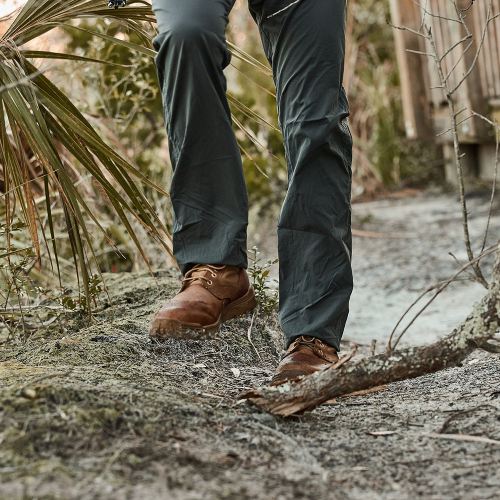 A person strolls along a forest path, wearing GORUCK's MACV-2 - Mid Top in Briar + Coyote with green pants, surrounded by lush plants and branches.