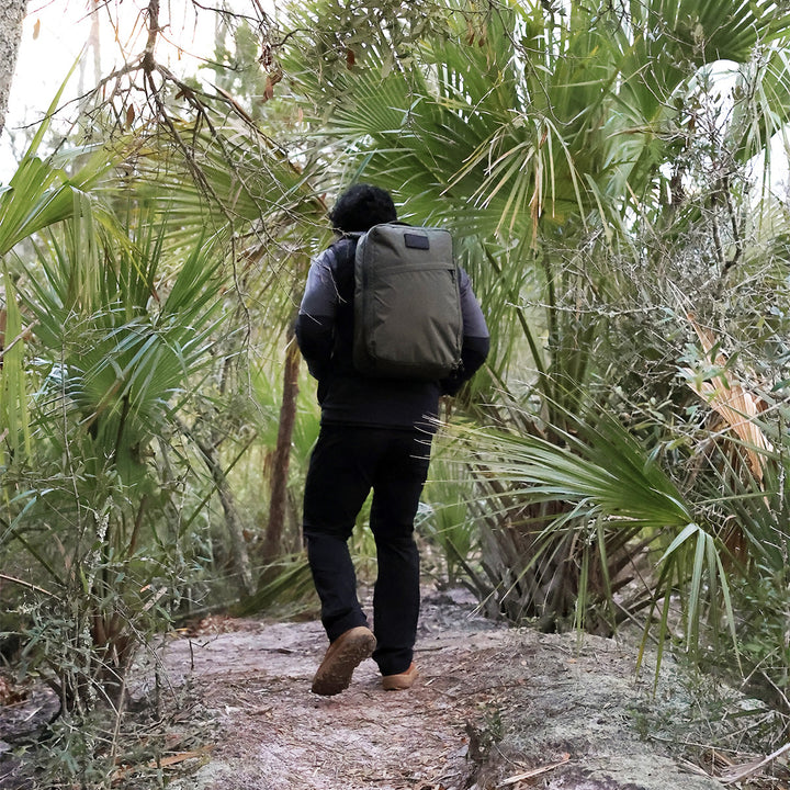 A hiker with a large backpack treks through the forest trail, wearing GORUCK's MACV-2 Mid Top boots in Briar + Coyote, featuring durable construction and an aggressive triple compound outsole, surrounded by lush green plants and towering palm trees.