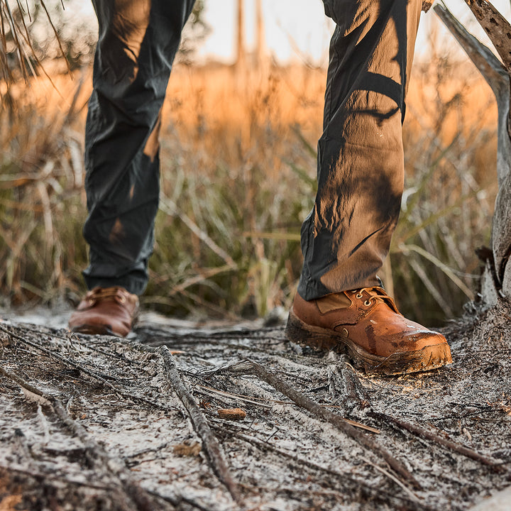 GORUCK's MACV-2 Mid Top in Briar + Coyote, with its aggressive triple compound outsole, provides superior grip and stability while hiking through a muddy wooded area at sunset.