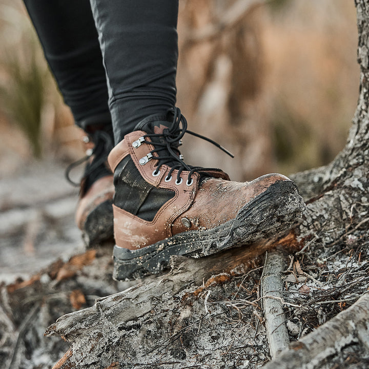 Close-up of GORUCK MACV-2 - Mid Top - Chestnut + Black hiking boots with an aggressive outsole on a rugged trail filled with roots and dirt, worn by someone in black pants.