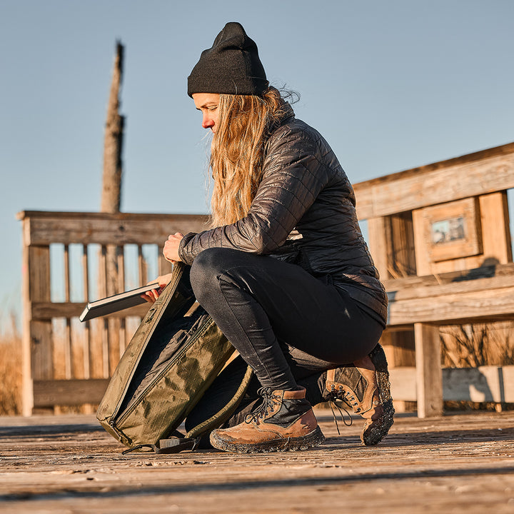 A woman in outdoor gear kneels on a wooden deck, packing her green backpack. She wears a beanie and jacket; beside her is the GORUCK MACV-2 - Mid Top in Chestnut + Black, its rugged outsole ideal for any trail.