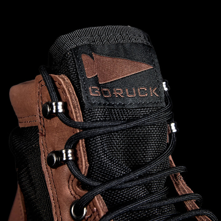 Close-up of the MACV-2 - Mid Top in Chestnut + Black with GORUCK and an arrowhead logo on the tongue against a black background, highlighting its aggressive outsole.