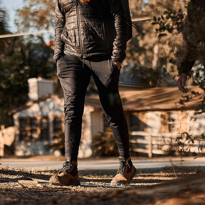 A person in a jacket and joggers strolls a sunny path with trees and a house nearby, confidently sporting GORUCK's MACV-2 - Mid Top - Chestnut + Black jungle boots featuring an aggressive outsole.