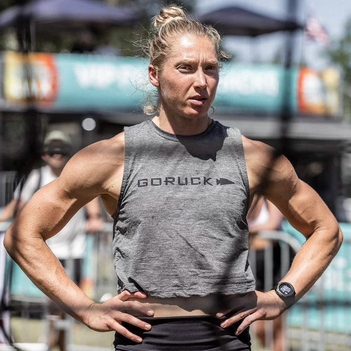 An athletic individual with a focused expression stands outdoors, wearing the GORUCK Women's Performance Tank - ToughMesh and a watch, hands on hips. The ToughMesh™ fabric enhances performance as blurred figures, trees, and a sign with visible text form the backdrop.