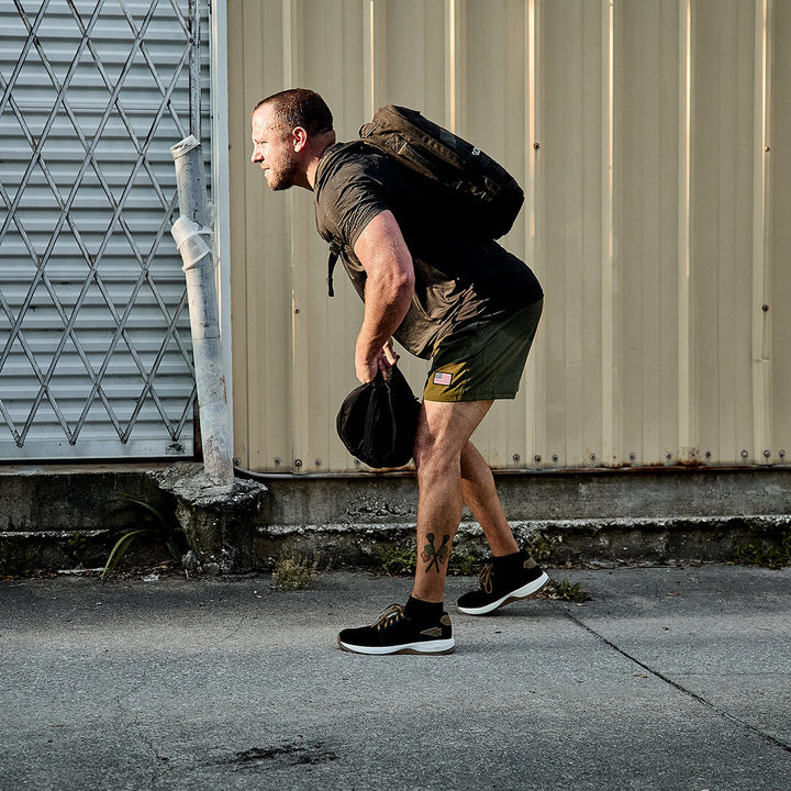 A man wearing a GORUCK backpack made from CORDURA Ballistic Nylon and dressed in casual athletic attire, including the Men's Ballistic Trainers in Black + White with Coyote Reflective Spearhead, leans forward as he walks on a concrete path beside a beige corrugated wall and chain link fence. The sun casts striking shadows, accentuating his focused expression and dynamic posture.