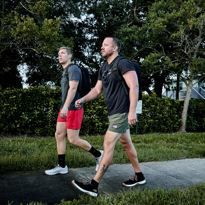 Two men walk leisurely along the sidewalk near a grassy area and trees, dressed in casual athletic attire and backpacks crafted from resilient CORDURA Ballistic Nylon. One man is wearing a gray shirt paired with red shorts, while the other sports a black shirt with green shorts. They are surrounded by vibrant bushes and trees, their attire complemented by GORUCK Men's Ballistic Trainers – Black + White with Coyote Reflective Spearhead accents.