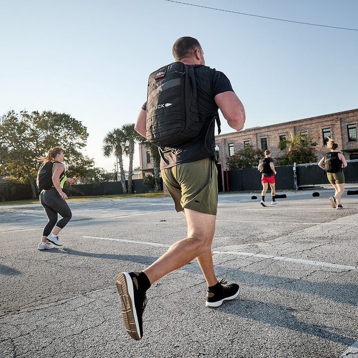 A group of people in athletic wear runs outdoors in a parking lot during the day. Prominent in the foreground is a man wearing Men's Ballistic Trainers by GORUCK in Black + White with Coyote Reflective Spearhead and carrying a black backpack made from CORDURA Ballistic Nylon. Trees and a building provide the backdrop for this display of functional fitness.