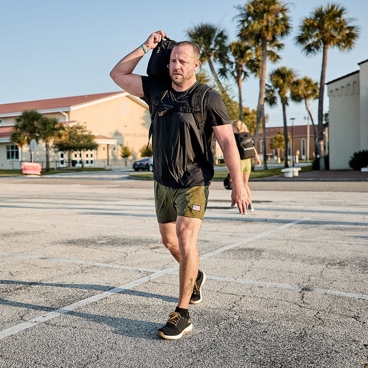 A man dressed in athletic gear, featuring GORUCK's Men's Ballistic Trainers in Black + White with Coyote Reflective Spearhead, walks across a parking lot carrying a large sandbag on his shoulder. He wears a black shirt and green shorts made from CORDURA Ballistic Nylon. Palm trees and buildings frame the scene under a clear blue sky.