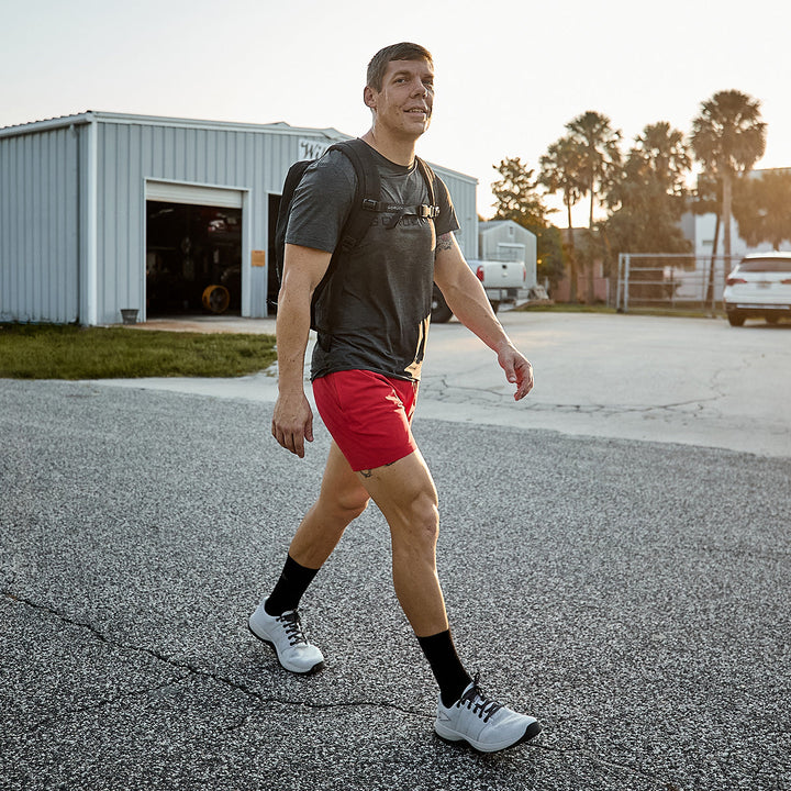 A man strolls along a paved road, clad in a gray T-shirt and red shorts, with his GORUCK Men's Ballistic Trainers in Lunar Rock and Charcoal with a Silver Reflective Spearhead providing functional fitness support. His backpack, crafted from CORDURA® Ballistic Nylon, hangs securely on his shoulders. As the sun dips low, a metal building and trees frame his path.