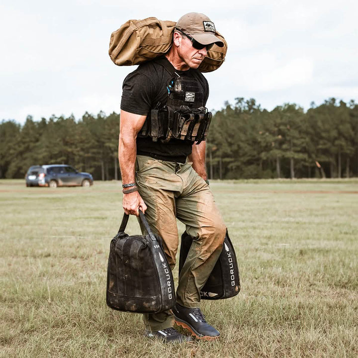 A person wearing GORUCK's Men’s Challenge Pants, crafted from Lightweight ToughDry® fabric featuring abrasion resistance and fast-drying capabilities with articulated knees, carries two weighted bags and a backpack while walking across a grassy field. In the background, there's a tree line and a parked car. They also sport a cap and sunglasses.