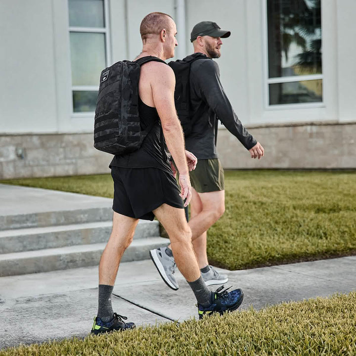 Two men, embodying the spirit of GORUCK's Rough Runner in Midnight Frogskin + Acid Lime, walk along the sidewalk in athletic gear and backpacks, likely discussing their road-to-trail outsoles and comfortable gradient density EVA midsoles near a building.