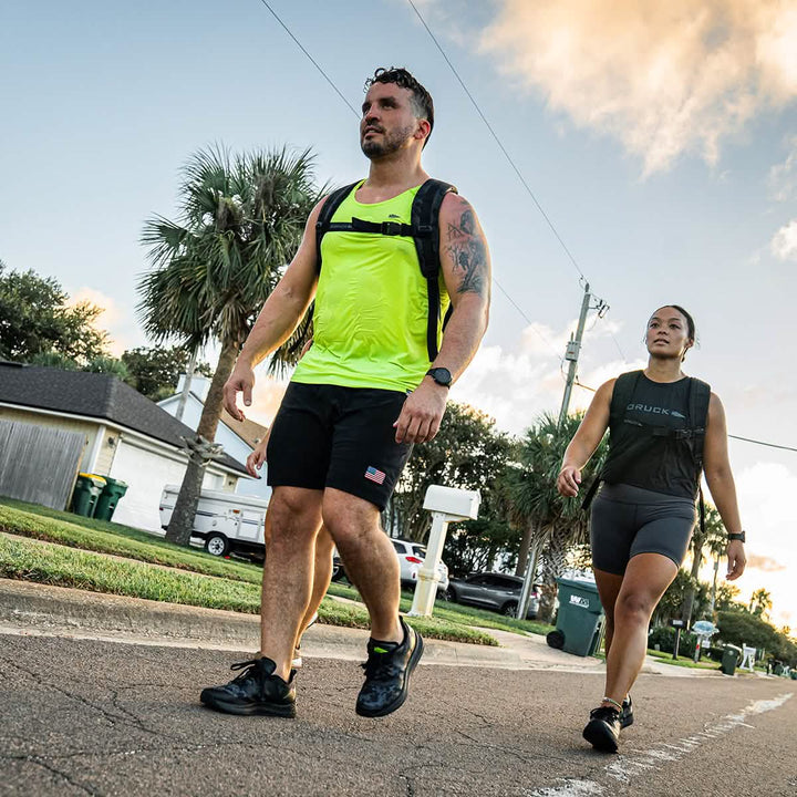 Two GORUCK Rough Runners, equipped with athletic gear and backpacks, walk along a suburban street filled with palm trees and houses. Their shoes, the Midnight Frogskin + Acid Lime edition, boast a Gradient Density EVA Midsole and a Versatile Road-to-Trail Outsole for diverse terrain.