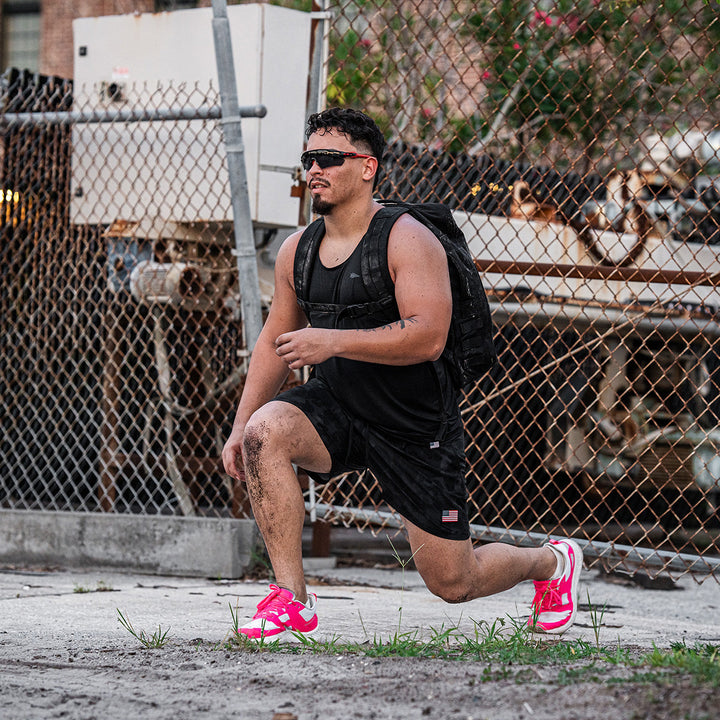 Near a chain-link fence, a person lunges outdoors wearing the Rough Runner - Hot Pink backpack and ultra-supportive running shoes by GORUCK, featuring a versatile road-to-trail outsole, complemented by sunglasses.