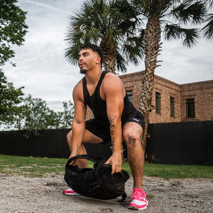 A man, wearing the ultra-supportive GORUCK Rough Runner in Hot Pink, lifts a heavy backpack outdoors near a brick building and palm trees. The versatile road-to-trail outsole suggests he's prepared for any adventure the day might bring.