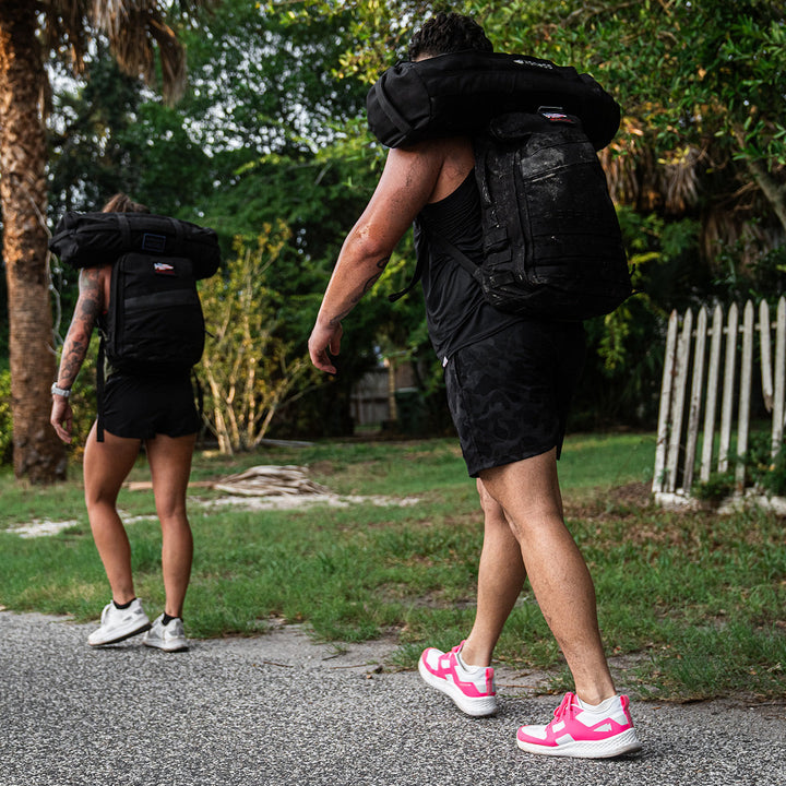 Two people walk along a road, carrying weighted sandbags on their shoulders, surrounded by lush greenery. They step confidently in GORUCK's Rough Runner - Hot Pink shoes, which offer ultra-supportive stability with a versatile road-to-trail outsole every step of the way.