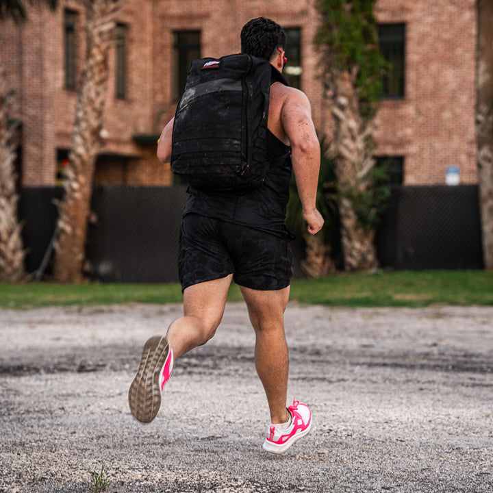 A person wearing the Rough Runner - Hot Pink from GORUCK dashes along a gravel path, their ultra-supportive shoes providing grip near palm trees and a brick building.