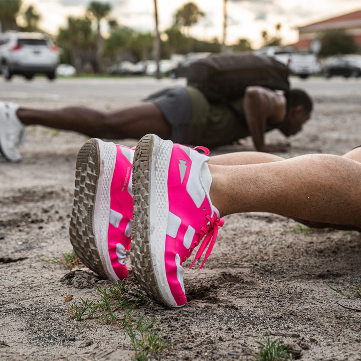 A person doing push-ups outdoors showcases their ultra-supportive Rough Runner - Hot Pink running shoes by GORUCK, featuring a versatile road-to-trail outsole and a striking pink and white design.