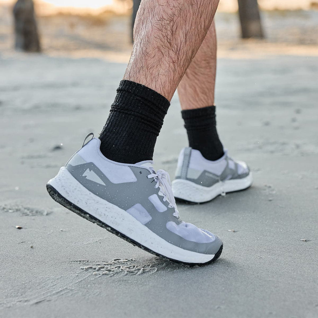 Close-up of a person strolling on a sandy beach wearing gray and white sneakers paired with GORUCK Merino Challenge Socks in black.