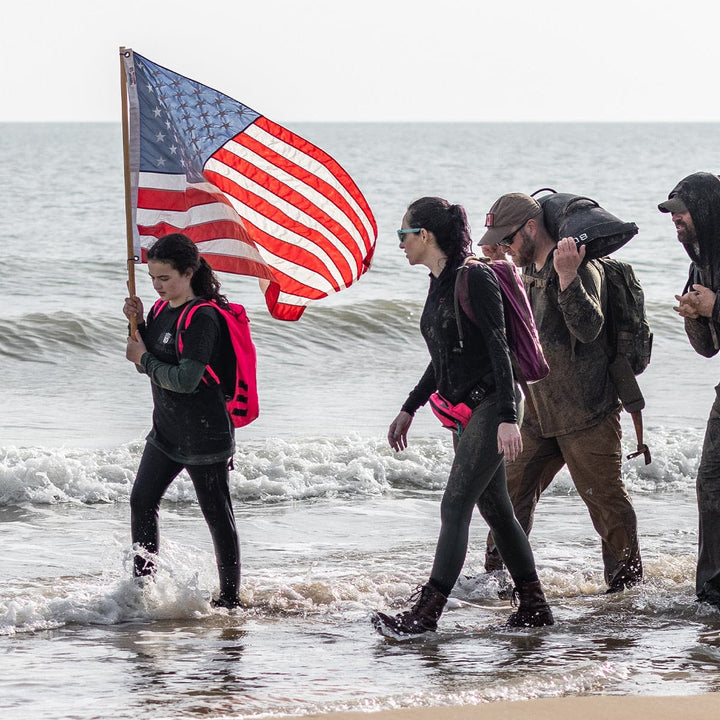 As people stroll along the beach in shallow water, they keep comfy wearing GORUCK's Merino Challenge Socks. One person carries an American flag while using a merino wool backpack that ensures durability for countless adventures, supported by the Scars Lifetime Guarantee.