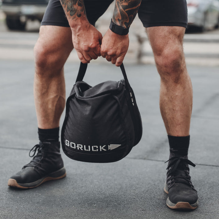 A person lifts a black sandbag outdoors, wearing GORUCK's sturdy Ballistic Trainers - Mid Top - Black + Gum with Black Reflective Spearhead sneakers designed for stability. The sandbag, crafted from durable CORDURA® Ballistic Nylon, promises enduring performance in any environment.