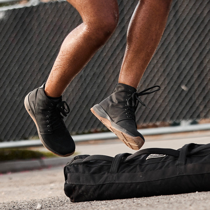 A person jumping mid-air in GORUCK's Ballistic Trainers - Mid Top - Black + Gum with a Black Reflective Spearhead, which showcase 3X Support and Stability, with a sleek black duffel bag crafted from CORDURA® Ballistic Nylon resting on the ground.