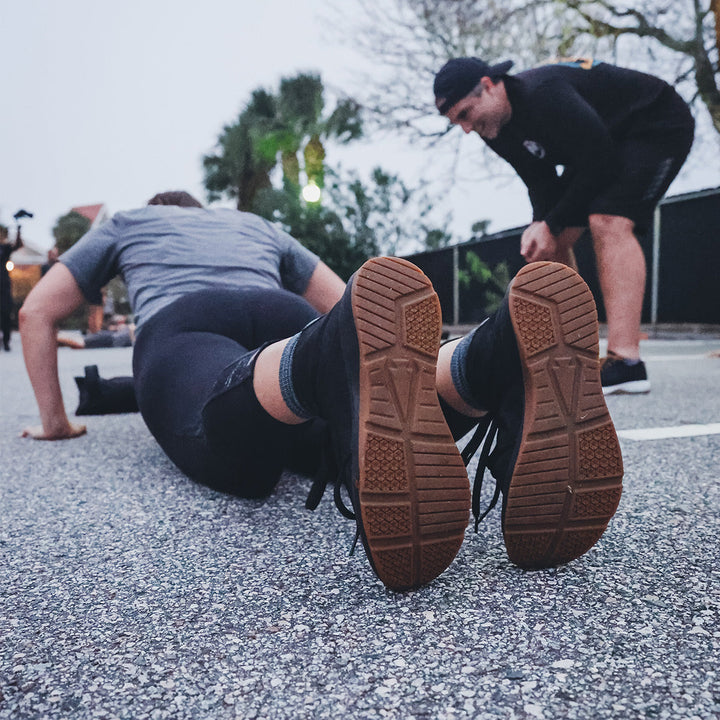 Two people exercising outdoors: one performs push-ups with optimal stability while the other, wearing GORUCK Ballistic Trainers - Mid Top in Black + Gum with a Black Reflective Spearhead, coaches or observes intently.