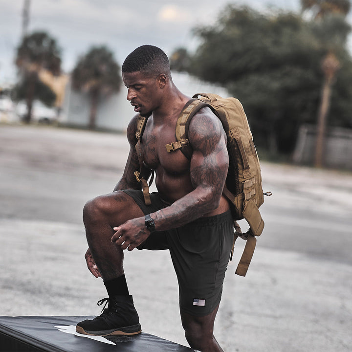 A man, wearing black shorts and exercising outdoors, demonstrates his stability as he steps onto a platform. He uses the GORUCK Ballistic Trainers - Mid Top in Black + Gum with Black Reflective Spearhead to support his rigorous workout.