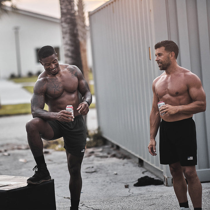 Two men wearing Ballistic Trainers from GORUCK are outdoors near a building with cans in hand. One of them is smiling and resting a foot on a crate, showcasing relaxed stability.