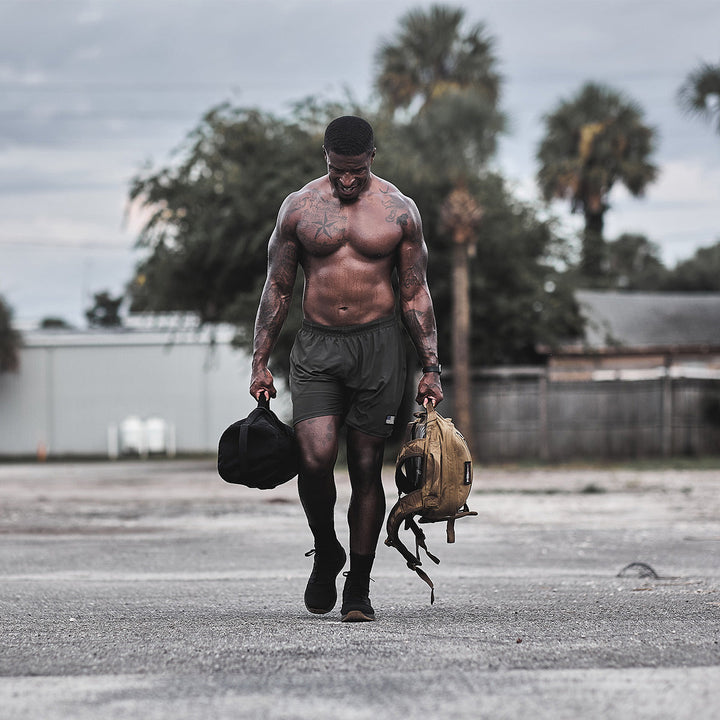 A muscular, tattooed man walks with stability outdoors in his GORUCK Ballistic Trainers - Mid Top in Black + Gum with a Black Reflective Spearhead, carrying a backpack and duffel bag made from CORDURA® Ballistic Nylon against a backdrop of trees.